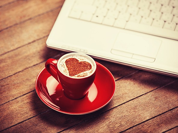 coffee in red cup with a love heart shape in the milk, on table next to a laptop